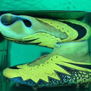 Gula Nike Hypervenom fotbollsskor - Säljer ett par gula Nike Hypervenom fotbollsskor med svart mönster och högt skaft. Skorna har snörning och en platt sula med dobbar, perfekt för spel på konst och naturligt gräs . Snygg design med svarta detaljer och Nike-logga på sulan.