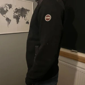 Svart jacka från Colmar med huva - Säljer en svart jacka från Colmar med huva och dragkedja. Jackan har diskret logotyp på ärmen och fickor. Perfekt för dig som vill ha en stilren och enkel jacka med sportig känsla.