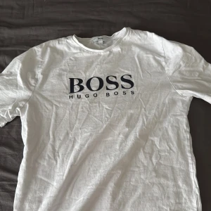 Vit t-shirt från Hugo Boss - Vit t-shirt från Hugo Boss med klassisk rund hals och stor svart logga på bröstet. Enkel och stilren design som passar perfekt till jeans eller shorts. Tillverkad i mjuk bomull för en bekväm känsla hela dagen. För 14 åriga ungdomar men passar som XXS