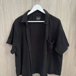 Svart kortärmad skjorta från Boohoo Man - Svart skjorta med kort ärm från Boohoo Man. Klassisk krage, knäppning framtill och en bröstficka. Skjortan har en avslappnad passform och är perfekt för en stilren look.