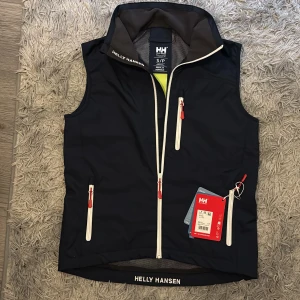 Marinblå väst från Helly Hansen (NY) - Snygg marinblå väst från Helly Hansen med vita dragkedjor och röda detaljer. Västen har ståkrage, tre fickor med dragkedja och logga på kragen samt nederkanten. Perfekt för lager-på-lager under kyliga dagar. Helt ny och aldrig använd.
