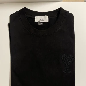 Svart t-shirt från Ami Paris - Säljer en svart t-shirt från Ami med det ikoniska broderade hjärtat med A på bröstet. T-shirten har rund halsringning och är i mjuk bomull. Perfekt för en stilren och enkel look.