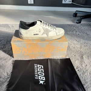 Vita sneakers från Golden Goose - Säljer ett par vita sneakers från Golden Goose med grå snörning, svart häl och den klassiska stjärnan på sidan. Skorna har platt sula och rund tå. Perfekta för dig som gillar stilrena och trendiga skor. Pris kan diskuteras vid snabb affär. Allt og medföljer