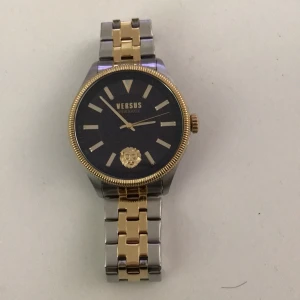 Versus Versace herrklocka i guld och silver - Stilren herrklocka från Versus Versace med svart urtavla, guldfärgade detaljer och lejonmotiv. Armbandet är i tvåtonad metall, guld och silver. Klockan har tydliga indexmarkeringar och klassisk design. Använd fåtal gånger nästan som ny pris kan diskuteras har även paket och allt