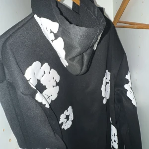 Svart hoodie med vita blommönster ifrån denim tears - Säljer en svart hoodie med stora vita blommönster tryckta över hela tröjan. Hoodien har lång ärm och en klassisk huva. Perfekt för dig som vill sticka ut med en cool och unik stil. Anledningen till att jag säljer denna är för att den var för stor för mig och den är helt ny