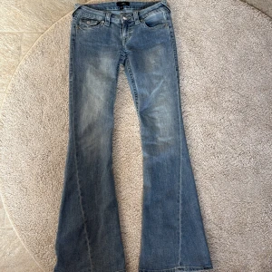 Blå bootcut jeans från True Religion - Säljer ett par klassiska blå bootcut jeans från True Religion med snygga fickdetaljer och logga bak. Jeansen har låg midja och utsvängda ben, samt dekorativa sömmar och knappar på bakfickorna.