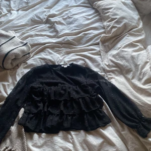 Svart volangblus från Gina Tricot - Säljer en svart blus från Gina Tricot med volanger framtill och små diskreta prickar i tyget. Blusen har långa ärmar och en lätt transparent känsla. Säljer den då den är för liten för mig 