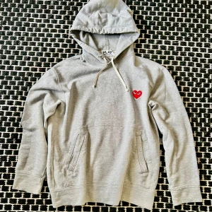 Grå hoodie från Comme des Garçons Play - Säljer en grå hoodie från Comme des Garçons Play med det ikoniska röda hjärtat broderat på bröstet. Tröjan har huva med dragsko och fickor framtill. Perfekt för en avslappnad och stilren look. Snall hole on the right sleeve