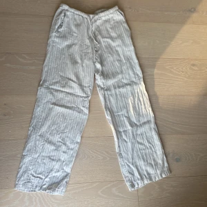 Ljusgråa randiga linnebyxor från Gina Tricot - Säljer ett par ljusgråa linnebyxor från Gina Tricot med vita ränder och dragsko i midjan. Byxorna har en avslappnad passform och raka ben, perfekta för varma dagar.
