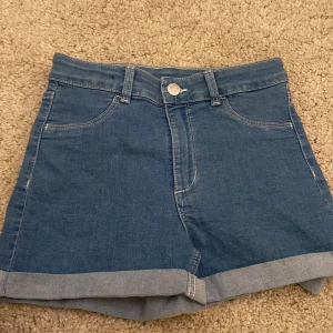 Blå jeansshorts med uppvik - Klassiska blå jeansshorts från &Denim med normal passform och uppvikta benslut. Shortsen har fem fickor och stängs med knapp och dragkedja. Perfekta för sommardagar och enkla att matcha med olika toppar.
