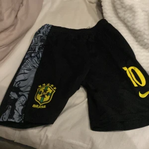 Svarta Brasilien fotbollsshorts med gula detaljer - Säljer ett par svarta fotbollsshorts från Nike med Brasilien-logga och nummer 10 i gult. Shortsen har en unik grå och svart mönstrad panel på sidorna. Perfekta för fotbollsträning eller match!