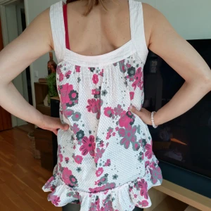 Vit blommig kortklänning med volang - Söt vit kortklänning med rosa och grå blommor samt små prickar. Klänningen har breda axelband, volang nertill och en lös passform. Perfekt för varma dagar och har en lekfull, somrig känsla.