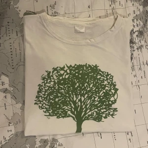 Vit t-shirt med grönt trädtryck - Vit t-shirt med ett stort grönt trädmotiv på framsidan. T-shirten har rund halsringning och är kortärmad. Naturinspirerade prints och enkel stil.