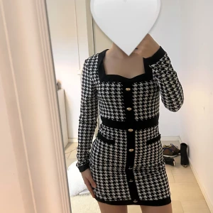 Svartvit rutig kortklänning med guldknappar - Säljer en snygg svartvit kortklänning med rutigt mönster och markerad midja. Klänningen har långa ärmar, dekorativa guldknappar framtill och svarta detaljer vid halsringning, midja och fickor. Perfekt för dig som gillar klassisk men trendig stil.