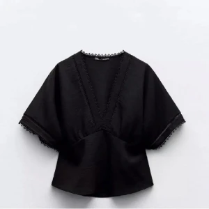 Svart blus från Zara - Svart blus med djup v-ringning och dekorativa spetsdetaljer längs halsringning och ärmslut. Modellen har korta, vida ärmar och en lös passform som ger en avslappnad men stilren look.