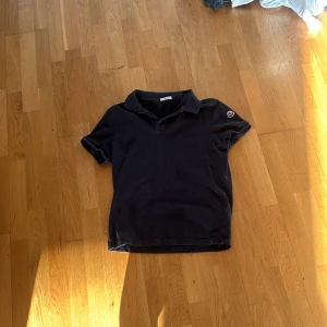 Mörkblå pikétröja från Moncler - Säljer en mörkblå pikétröja från Moncler med klassisk krage och korta ärmar. Moncler-logga på ena ärmen och broderad Moncler-text i rött och vitt på ryggen. Perfekt för en stilren och sportig look.