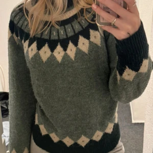 Grön stickad tröja med mönster från ONLY Studio - Mysig grön stickad tröja från ONLY Studio med beige och mörkgrönt geometriskt mönster runt halsen, ärmslut och nederkant. Tröjan har lång ärm och hög hals, perfekt för kyligare dagar. Så varm och skönt material🥹🥹🙌🏼