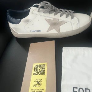 Golden Goose Superstar sneakers med grå stjärna - Golden Goose Superstar sneakers i vitt skinn med grå mockadetaljer och ikonisk stjärna på sidan. Skorna har mörkblå hälkappa, grå snörning och platt sula. Perfekta för dig som gillar en stilren men ändå unik look.