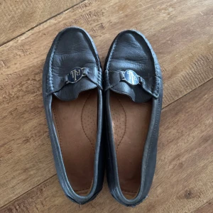 Svarta loafers från Ralph Lauren - Klassiska svarta loafers från Ralph Lauren med RL-logga i silver framtill och röd RL-detalj på hälen. Skorna är i mjukt skinn och har en platt sula samt rund tå. Perfekta för en stilren och avslappnad look.
