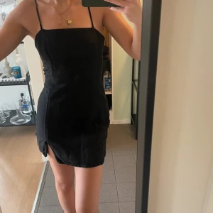 Svart kort klänning med snörning i ryggen - Säljer en svart kort klänning med smala axelband och slits på sidan. Klänningen har öppen rygg med snygg snörningsdetalj och figurnära passform. Perfekt för dig som vill ha en stilren och trendig look.