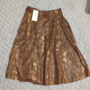 Brun kjol med ormmönster - Säljer en brun kjol med glansig finish och ormmönster. Kjolen har en lätt utsvängd form och går till knäna. Perfekt för dig som vill sticka ut med ett djurmönster i garderoben.