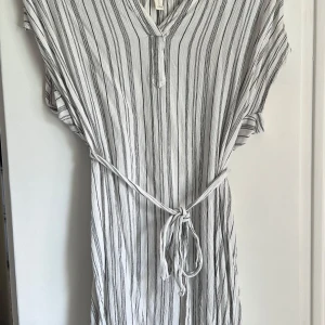 Vit och svart randig klänning från H&M L.O.G.G. - Säljer en luftig vit klänning med svarta ränder från H&M L.O.G.G. Klänningen har v-ringning, korta ärmar och ett knytband i midjan för en snygg passform. Perfekt för varma dagar! Passar även 34-38