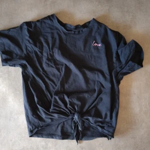 Svart t-shirt med knyt och broderad text - Svart t-shirt med kort ärm och knytning framtill. Liten broderad text 'love' i rosa på bröstet. Perfekt för en avslappnad och trendig stil.
