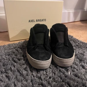 Svarta sneakers från Axel Arigato - Säljer ett par svarta sneakers från Axel Arigato med vit platt sula. Skorna är i mocka och har en stilren, minimalistisk design utan snörning. Perfekta för dig som gillar enkel och snygg streetstil. Skorna har några defekter men är annars i bra skick. Orginalbox och dustbag kommer även med i paketet