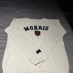 Vit stickad tröja från Morris - Säljer en vit stickad tröja från Morris med broderad logga och emblem på bröstet. Tröjan har lång ärm och har en klassisk, ribbad struktur. Perfekt för en stilren och avslappnad look. Öppen för prisförslag!