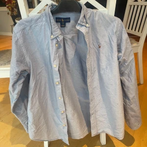 Ljusblå skjorta från Ralph Lauren - Klassisk ljusblå skjorta från Ralph Lauren med button-down krage och broderad logga på bröstet. Skjortan har vita knappar och lång ärm, perfekt för en stilren look.