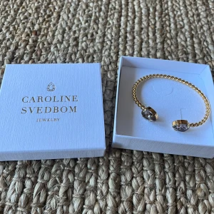 Guldfärgat armband med kristaller från Caroline Svedbom - Helt oanvänd! Säljer ett elegant armband från Caroline Svedbom i guldfärgad metall med två stora, gnistrande kristaller i färgen greige. Armbandet har en tvinnad design och är öppet framtill för enkel påtagning. Perfekt för att lyfta din look! Nypris 795kr.