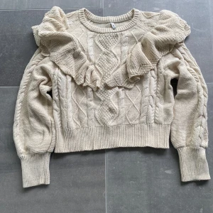 Beige stickad tröja med volang - Säljer en beige stickad tröja med volangdetaljer över axlarna och kabelstickat mönster framtill. Tröjan har rund halsringning, långa ärmar och ribbade muddar. Perfekt för dig som vill ha något både snyggt och bekvämt. OBS liten i storlek 