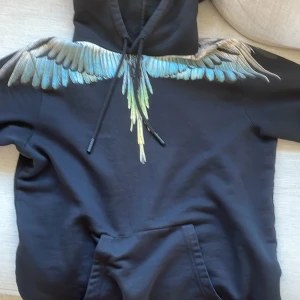 Svart hoodie med färgglada vingar från Marcelo Burlon - Säljer en svart hoodie från Marcelo Burlon med ikoniskt vingtryck i blått, grönt och beige över axlar och bröst. Tröjan har huva med dragsko och stor magficka framtill. Perfekt statement-plagg för dig som gillar unika detaljer.