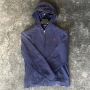 Marinblå hoodie från Polo Ralph Lauren - Säljer en marinblå hoodie från Polo Ralph Lauren i storlek 18-20 år som motsvarar S. Tröjan har dragkedja framtill, huva och fickor samt den klassiska röda loggan broderad på bröstet. Perfekt för en avslappnad stil. En defekt vid ena ärmkanten därav priset.