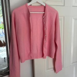 Rosa kofta från C Clothes - Söt rosa kofta från C Clothes i storlek S. Tillverkad i en mjuk blandning av lammull, nylon och angora. Koftan har långa ärmar och pärlknappar framtill som ger en extra fin detalj. Perfekt att slänga på sig när det är kyligt ute.