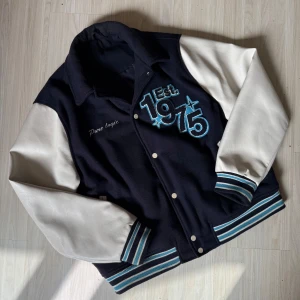 Svart och vit varsityjacka med broderi - Snygg varsityjacka i marinblå med vita ärmar och broderade detaljer. Framsidan har texten 'Est. 1975' och 'Pure Logic', baksidan har stort broderi med 'PURE LOGIC' och en megafon. Jackan har knappar och randiga muddar.