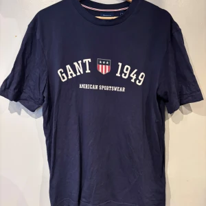Mörkblå t-shirt från Gant med tryck - Snygg mörkblå t-shirt från Gant med vit text och sköldtryck framtill. Klassisk rund halsringning och korta ärmar. Perfekt för en sportig och stilren look.