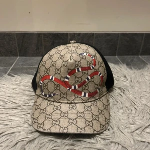 Beige keps från Gucci med ormtryck - Gucci keps i beige med klassiskt GG-mönster och en röd, vit och svart orm på framsidan. Baksidan är i svart mesh för extra ventilation. Märke och storlek står på insidan, 57 cm omkrets. Äkta