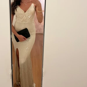 Champagnefärgad glittrig långklänning med slits - Säljer en elegant champagnefärgad långklänning med glittrigt material och v-ringad omlott-topp. Klänningen har en hög slits framtill och smala axelband. Perfekt för bal eller festkvällar.