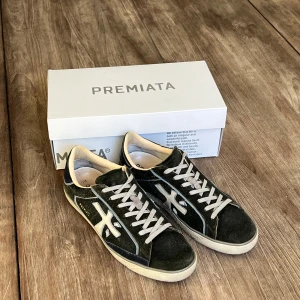 Premiata sneakers i mörkgrön mocka - Snygga sneakers från Premiata i mörkgrön mocka med vita detaljer och snörning. Skorna har en klassisk rund tå och platt sula, samt märkets logga på sidan. Perfekta för dig som gillar stilrena och bekväma skor. Storlek 44😁