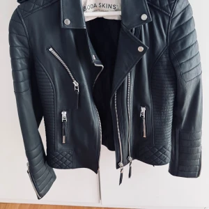 Svart skinnjacka från Boda Skins - Säljer en svart skinnjacka från Boda Skins med coola quiltade detaljer, flera dragkedjor och bältesdetaljer vid midjan. Jackan har lång ärm och klassisk bikerstil. Perfekt för dig som vill ha en edgy look. Storlek 34/36
