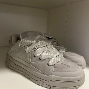 Grå sneakers (oanvända) - Säljer ett par stilrena grå sneakers med breda snören och chunky sula. Skorna har en modern design med paneler i syntetmaterial och mockaliknande detaljer. Perfekta för dig som gillar minimalistisk stil.