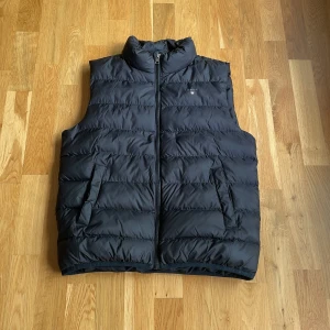 Svart dunväst från GANT - Säljer en svart dunväst från GANT med hög krage och dragkedja framtill. Västen har två stora fickor på sidorna samt en innerficka. Storleken är 170 cm men motsvarar ungefär Xs-S. Priset är inte hugget i sten!