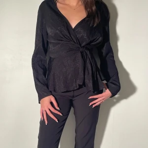 Svart omlottblus med knyt - Säljer en svart omlottblus med knyt i midjan och lång ärm. Blusen har en lätt glansig yta och djup v-ringning, perfekt att styla med både jeans och kostymbyxor. Snygg och enkel att matcha till olika looks.