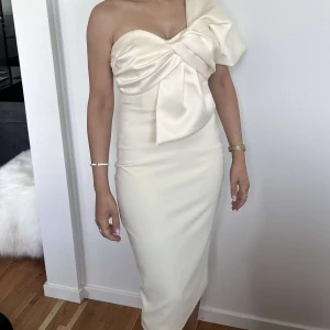 Champagnefärgad/beige one shoulder-klänning med rosettdetalj - Säljer en elegant champagnefärgad klänning med one shoulder och stor rosettdetalj framtill. Klänningen är figurnära och har en stilren, lång siluett. Perfekt för speciella tillfällen som student eller examenfirande💗Storlek 34/36. Använd i några timmar. Pris 500kr