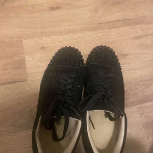 Svarta sneakers med nitar och röd sula - Svarta sneakers med snörning och coola svarta nitar på tån. Skorna har en tydlig röd sula undertill och är tillverkade i mocka. Perfekta för dig som vill sticka ut med din stil.