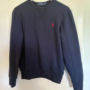 Mörkblå sweatshirt från Polo Ralph Lauren - En klassisk mörkblå sweatshirt från Polo Ralph Lauren med rund halsringning och röd broderad logga på bröstet. Tröjan har långa ärmar och ribbade muddar vid ärmslut och nederkant. Pris kan diskuteras!