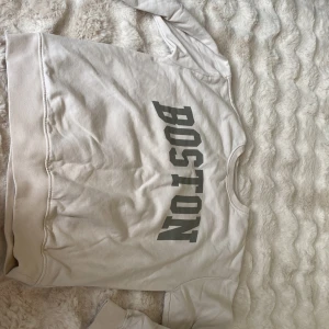 Vit sweatshirt med BOSTON-tryck - Vit sweatshirt med grått BOSTON-tryck på bröstet. Tröjan har rund halsringning, långa ärmar och ribbade muddar vid ärmslut och nederkant. Perfekt för en avslappnad och sportig stil.