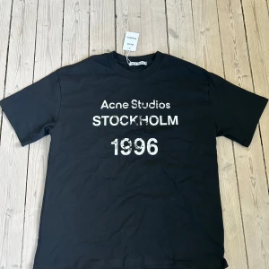 Svart t-shirt från Acne Studios med tryck - Svart t-shirt från Acne Studios med vitt tryck på bröstet: 'Acne Studios STOCKHOLM 1996'. Klassisk passform och rund hals. Passar även L