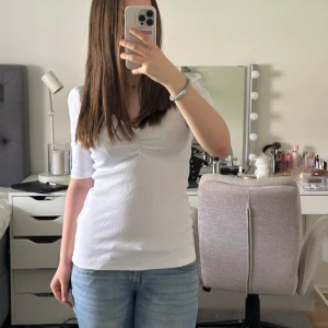 Vit ribbad topp med puffärm och v-ringning - Säljer en vit ribbad topp med kort puffärm och snygg v-ringning. Toppen har en rynkad detalj vid bysten och är figurnära i modellen. Perfekt att matcha med jeans eller kjol.